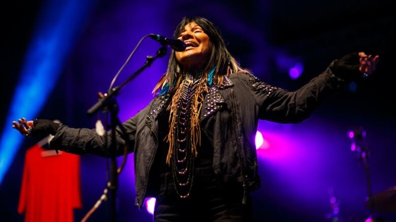 Buffy Sainte-Marie on stage sings into a microphone with her eyes closed and her arms outstretched. Blue and purple stage lights colour the background and a red dress also hangs in the background. 