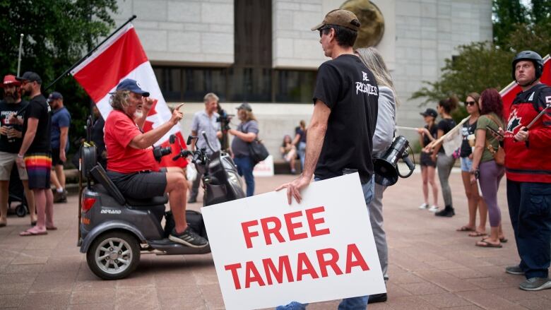 A man on a mobility scooter drives past a younger man in a black T-shirt holding a sign that reads 