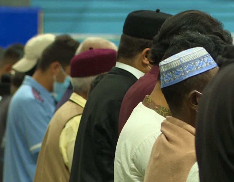 Men stand in a row, some of them wearing Muslim caps. Only the side of the men's heads can be seen.