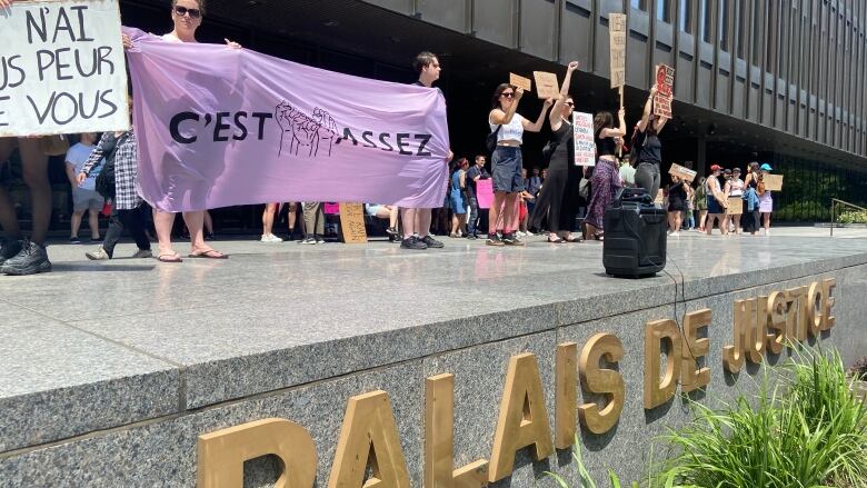 A group of dozens of protestors, many women, gather outside a courthouse with a sign reading Cest assez, French for Its enough.