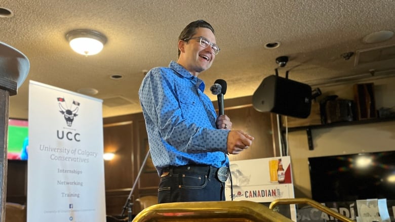 A smiling bespectacled politician dressed in a western shirt speaks into a microphone.