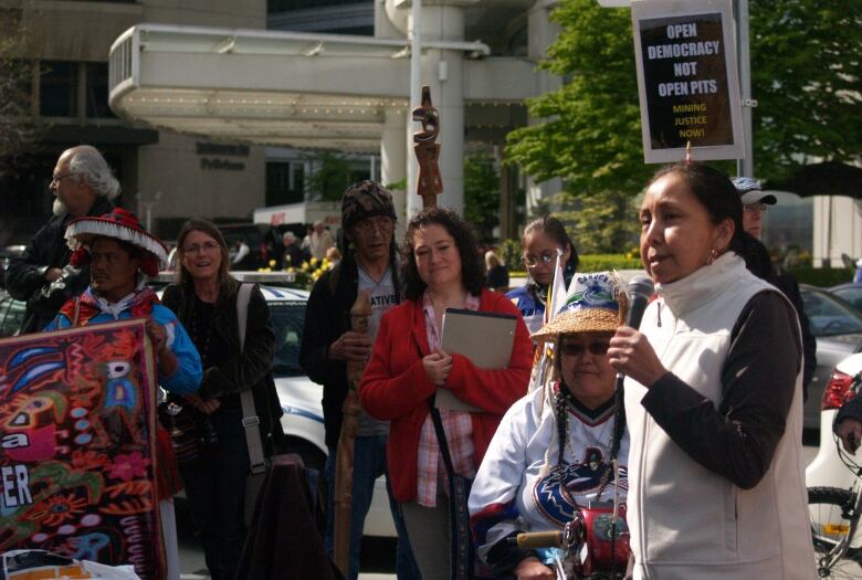 Kat Norris, founder of the Indigenous Action Movement and a member of Lyackson First Nation, holds a microphone and speaks at an anti-mining rally in Vancouver, B.C. in 2011. A crowd looks on as she speaks, and behind her is a sign that reads, 