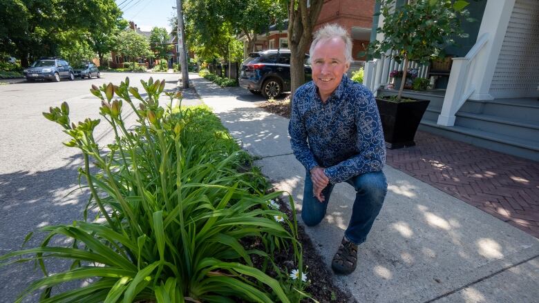 A man kneels and smiles in front of a garden planted in the narrow verge between a street and a sidewalk.