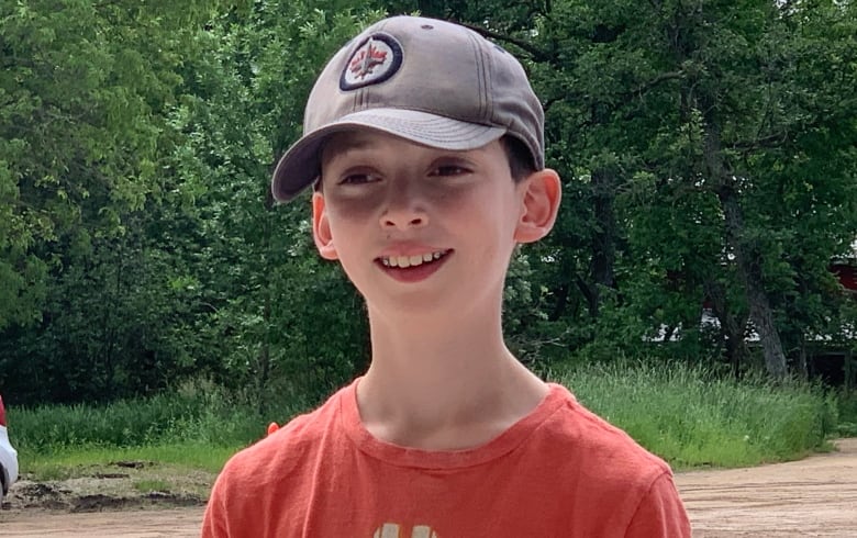 A boy wearing a Winnipeg Jets ballcap and an orange shirt is smiling in front of a gravel path and greenery.