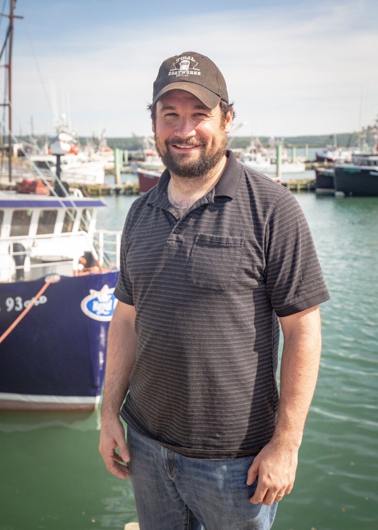 A man with a beard and ball cap stands in front of a harbour.