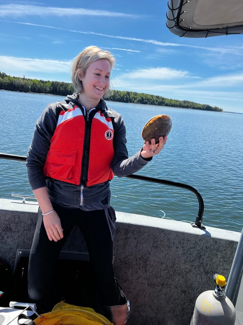 A smiling woman wearing a life jacket in a boat handles an Atlantic sea scallop. 