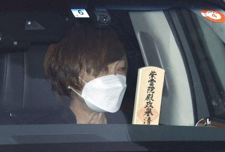 A closeup of a brown-haired woman passenger in a car is shown. She is carrying a fan with Japanese writing.