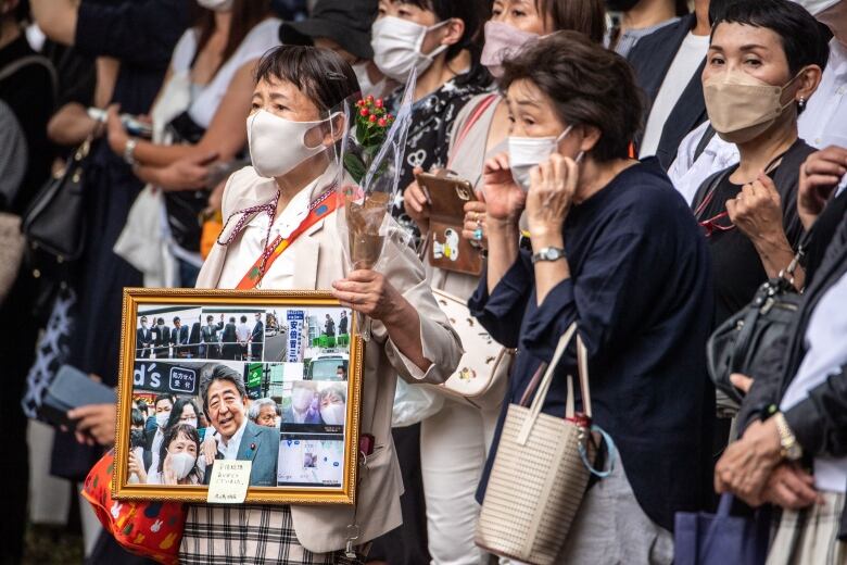 Several older women with COVID masks are shown in a crowd. One woman is holding a large frame with a photo collage inside it.