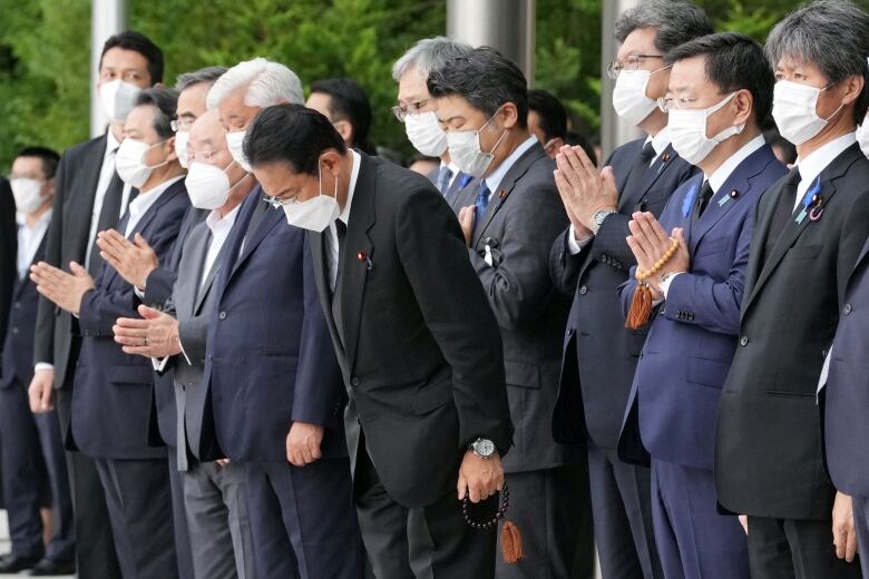 Several men in suits and COVID masks stand in a line. One of the men, stepping forward ahead of the others, bows.