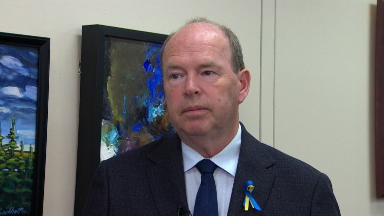 A man with thinning hair wearing a black jacket, white shirt and black tie. There's a colourful ribbon on his lapel.