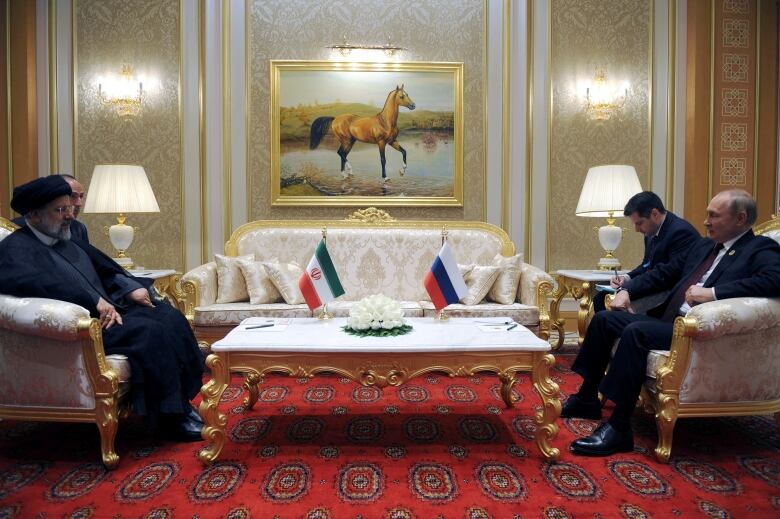 Two men sit on a couch across from two other men also on a couch. A low table with two flags on it is between them. A portrait of a horse hangs on the wall above them. 