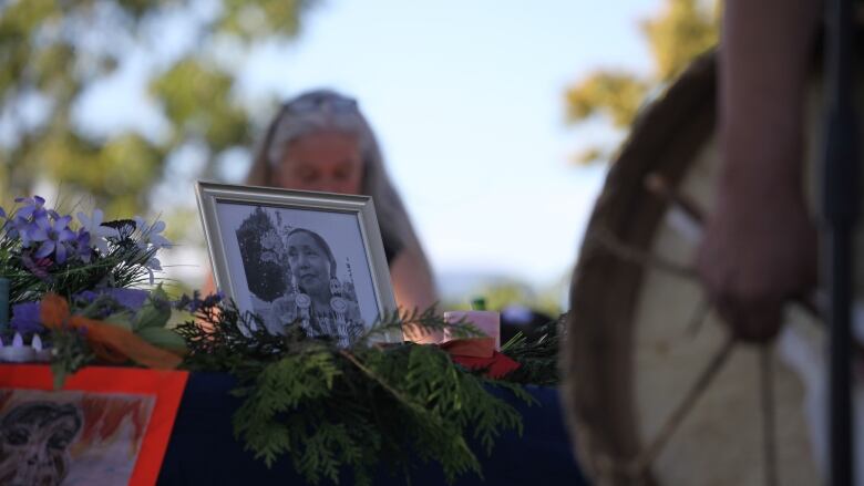 A photograph of Lyackson First Nation member Kat Norris is seen on a table with cedar, candles of sage at a memorial vigil in Grandview Park, Vancouver on Tuesday. A drum is held in the foreground as a woman pays respects from behind the table.