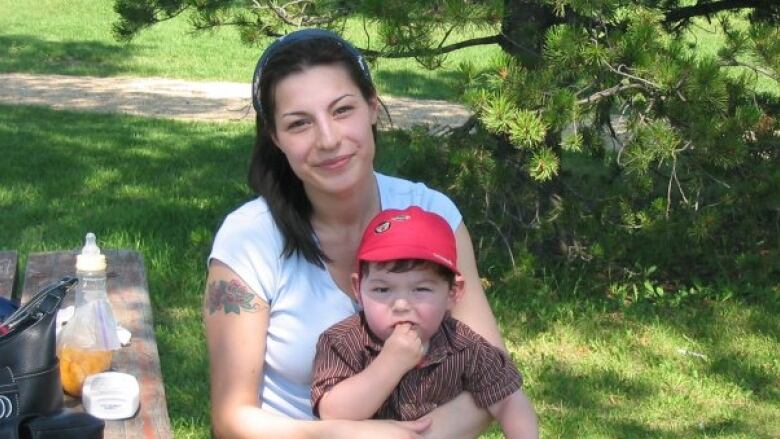 A smiling woman sits at a picnic table with her toddler son on her lap.