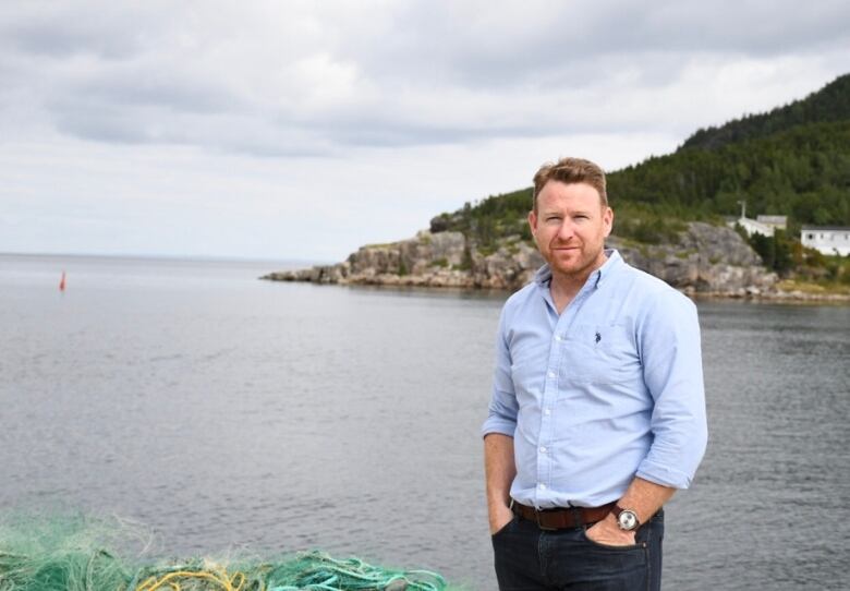 A man wearing an unbuttoned shirt and slacks stands in front of harbour and a hillside scene. The ocean is in the background. 