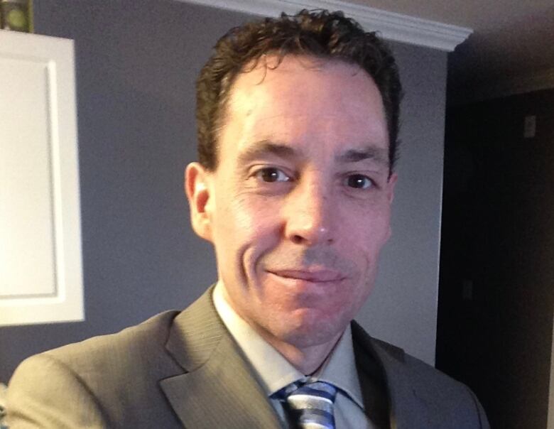 A man with short brown hair wearing a brown suit jacket with tie, smiling.