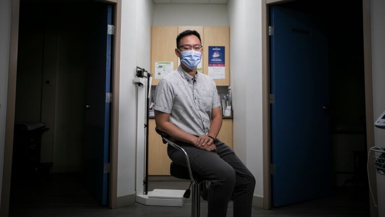 An Asian man with glasses sits in a doctor's office. He is wearing a grey shirt and a blue facemask.