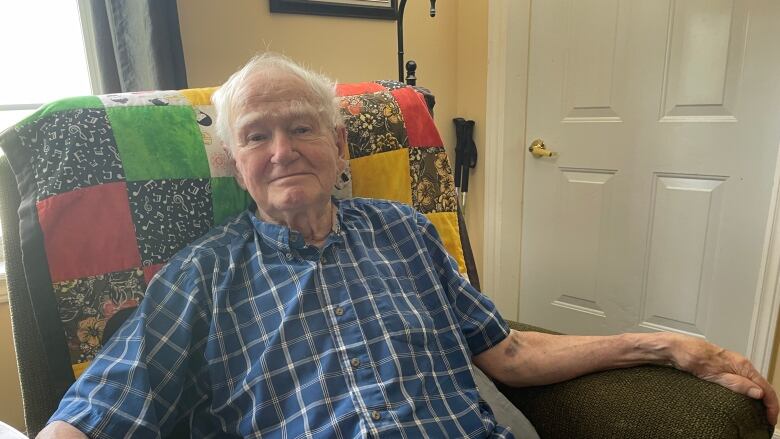 An older man sits in a recliner with a small smile.