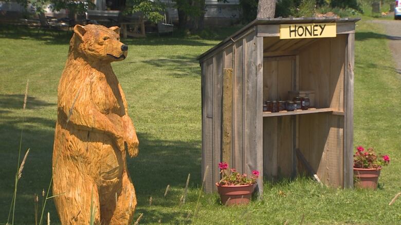 A carved wooden bear stands near a wood hut with a yellow sign that says Honey. Jars of honey sit on a shelf inside and geraniums in pots sit outside.