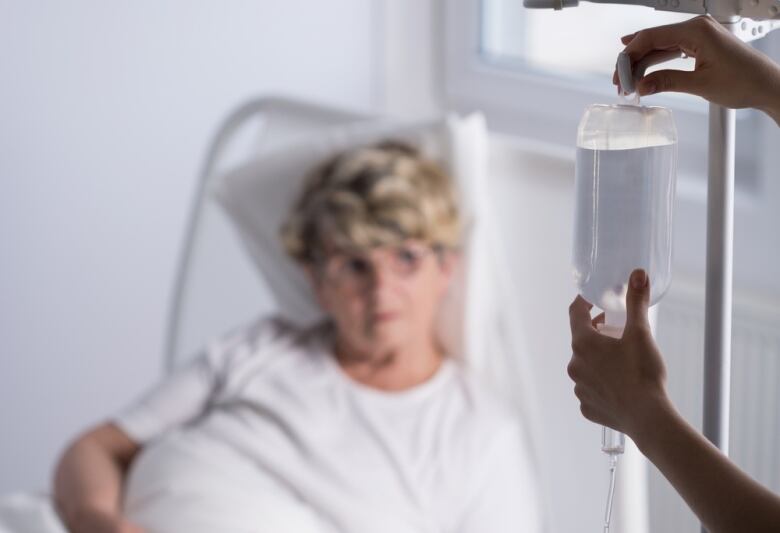 A person's hands are shown in the foreground, adjusting a fluid-filled bag for a patient lying in a hospital bed.