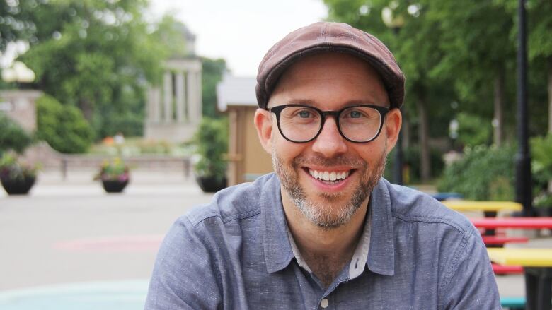 Portrait of a man with beard who is wearing glasses, a hat and a blue button-up shirt.