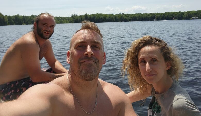 Three people smile sitting out in front of a lake. 