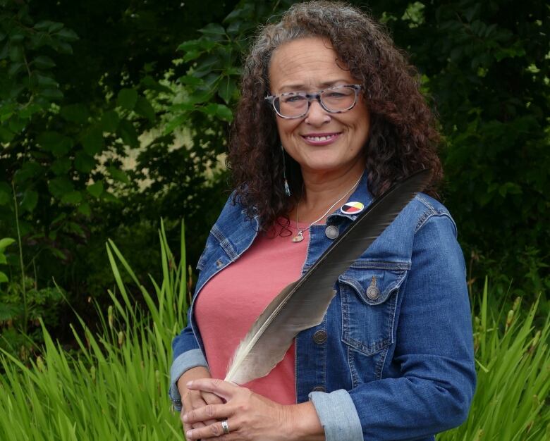 A woman in a jean jacket and glasses smiles at the camera, with trees in the background.