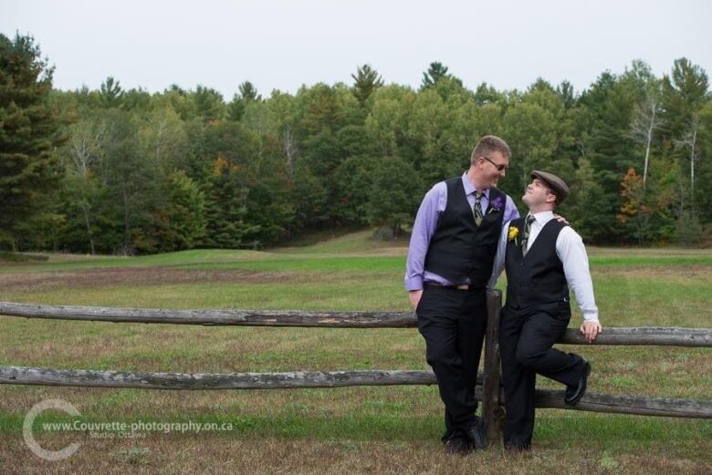 Two men lean on a wooden fence in a field of grass, looking at each other, with trees in the background.