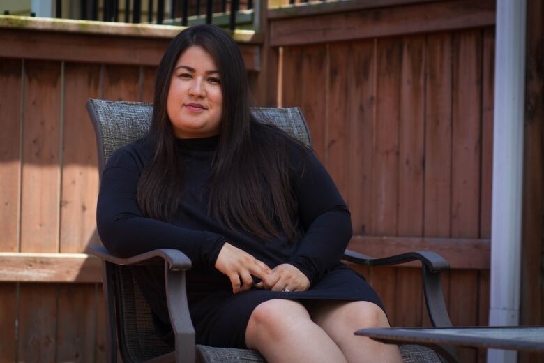 A woman with long brown hair sits on a patio chair and looks into the camera. There is a reddish brown fence behind her.