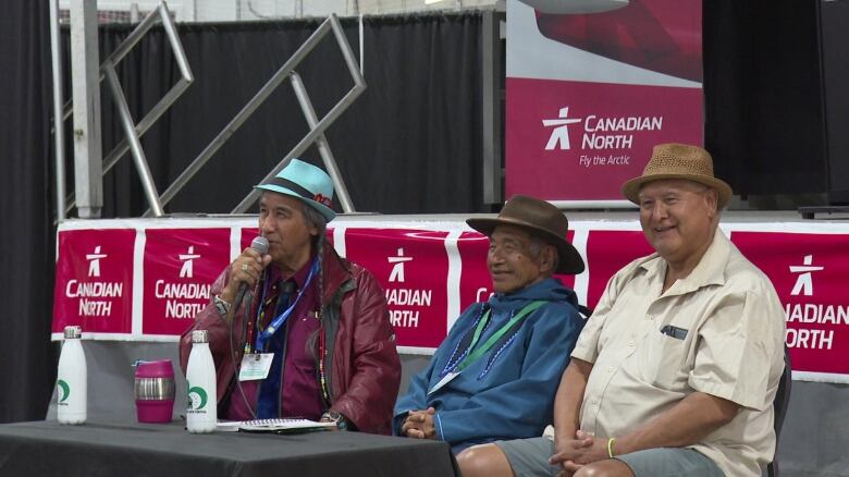 Three men, one holding a microphone and speaking, sit at a table.