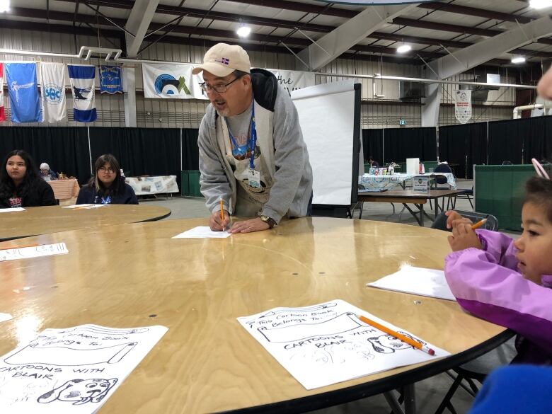 A man stands with a pencil poised over a sheet of paper, speaking with a circle of children around a table.