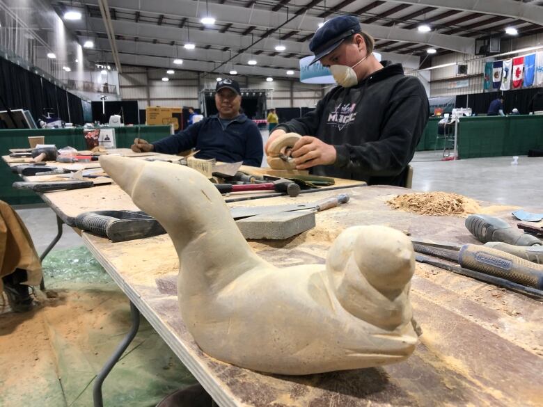 Two men sit at a table covered in dust, tools and carvings. One of them painstakingly carves a piece of soapstone.