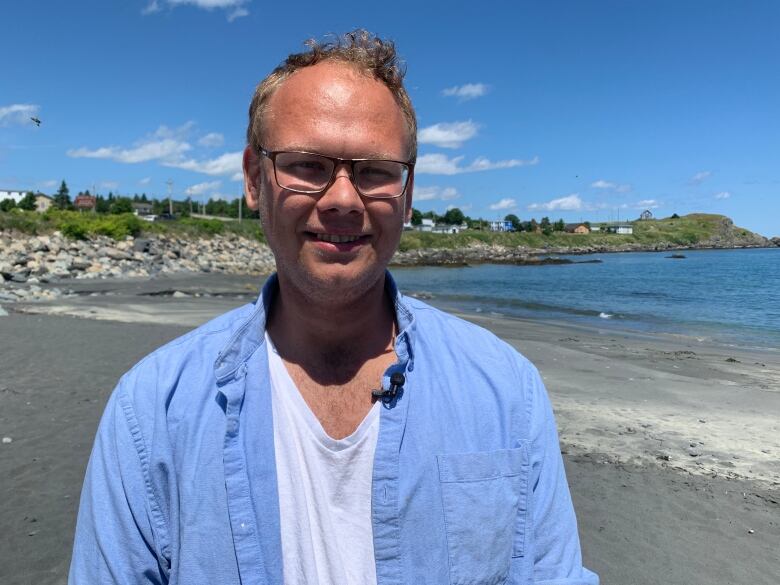 A man wearing glasses stands on a beach in the sunshine.