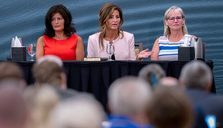 Three women are shown seated at a table in front of a crowd of people.