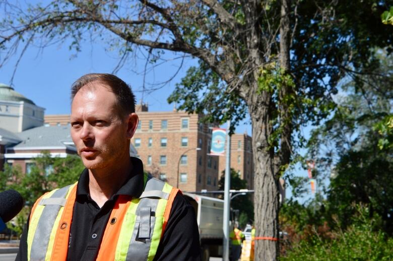 A man in a orange and yellow high-viz jacket stands infront of an elm tree in Victoria Park. 