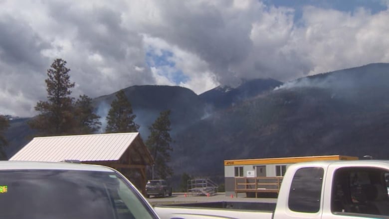 Plumes of smoke arise from two hills. Pickup trucks and containers are visible in the foreground.