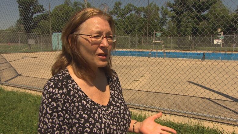 A woman with long hair and glasses is standing in front of a chain link fence surrounding a pool on a sunny day.