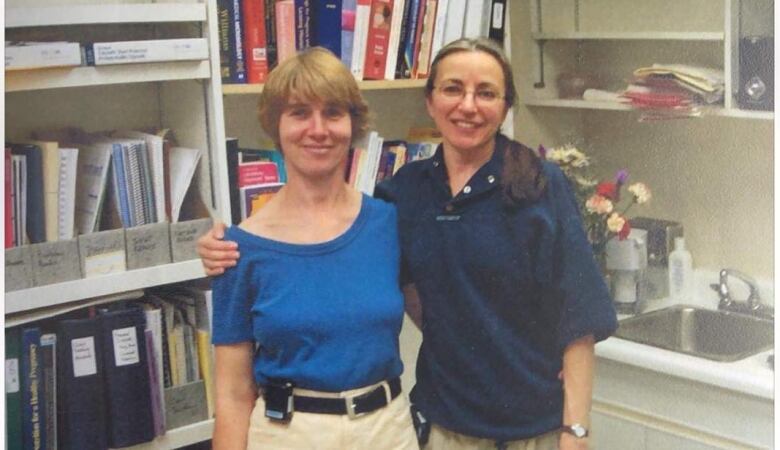 Two women with arms around each other's backs smile at the camera.