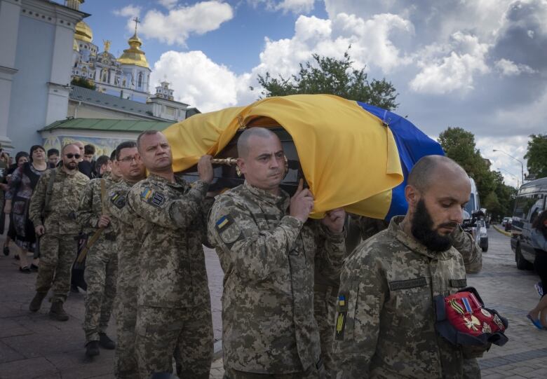 Men in military uniforms carry a coffin, draped in a blue and yellow flag, on their shoulders, in front of a white building with gold domes on top. 