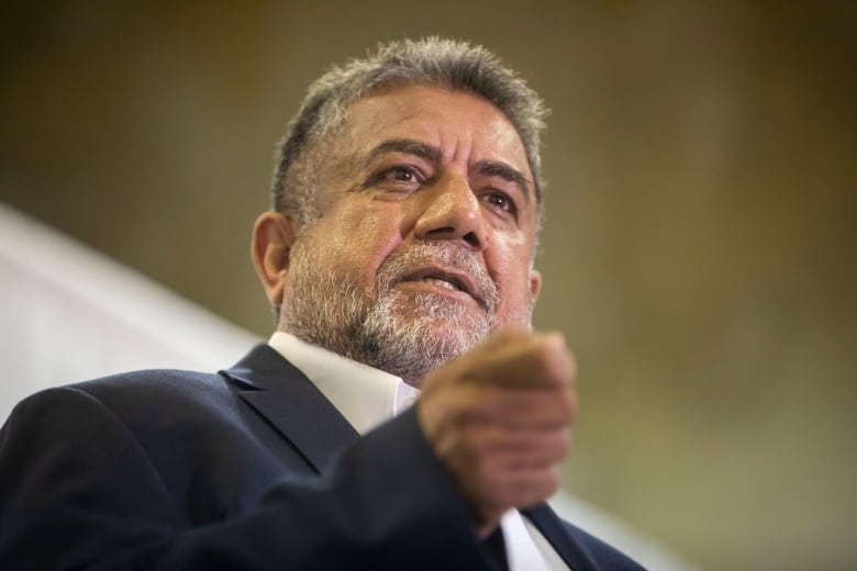 A South Asian man is pictured speaking with his hand outstretched.