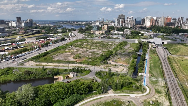 A dirt and grass field from above, with a river and city skyline in the background.