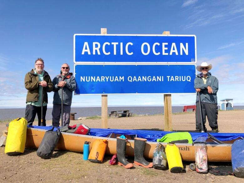 Three canoers stand on a beach with a sign reading 