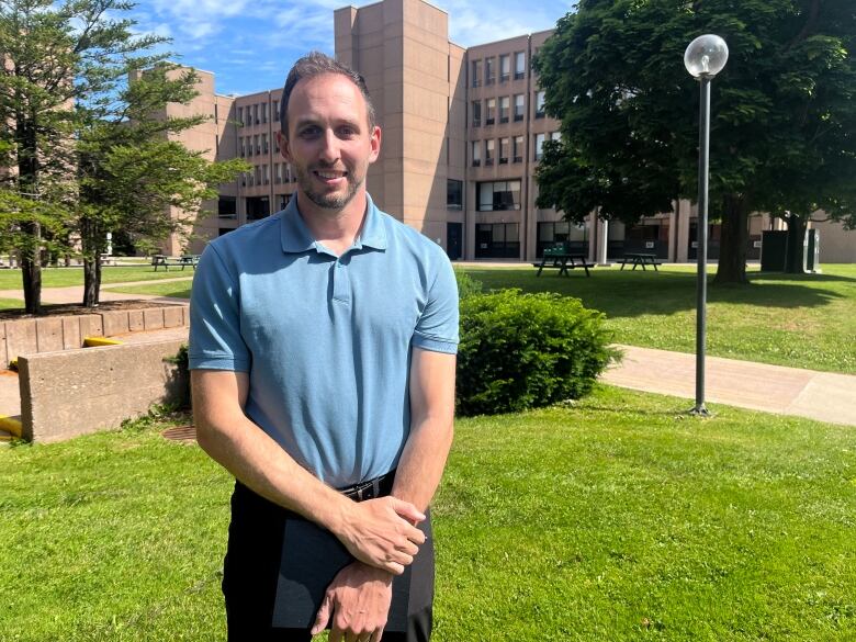 Man in blue polo shirt stands on lawn outside large brick building.