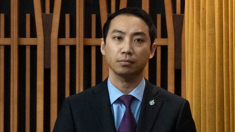 An Asian man with black hair wears a black suit, a light blue shirt and a purple tie. He has a solemn expression.