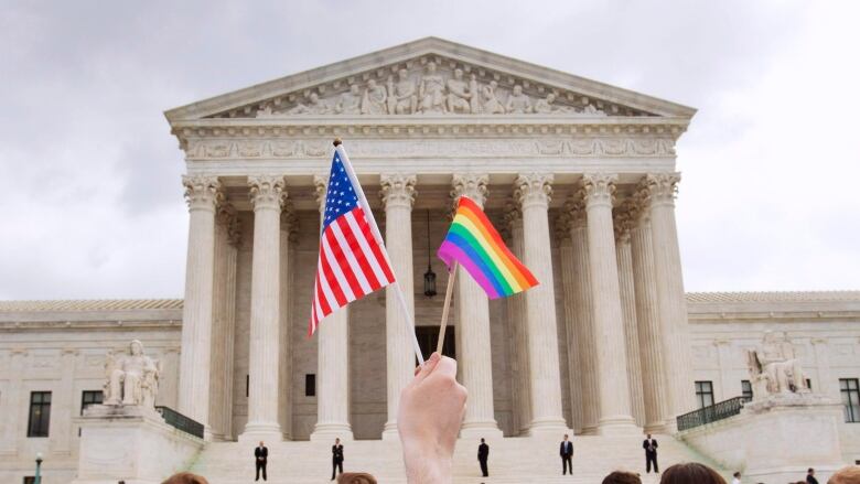 A hand holds up a U.S. flag and a rainbow Pride flag, above a crowd, in front of a building. 