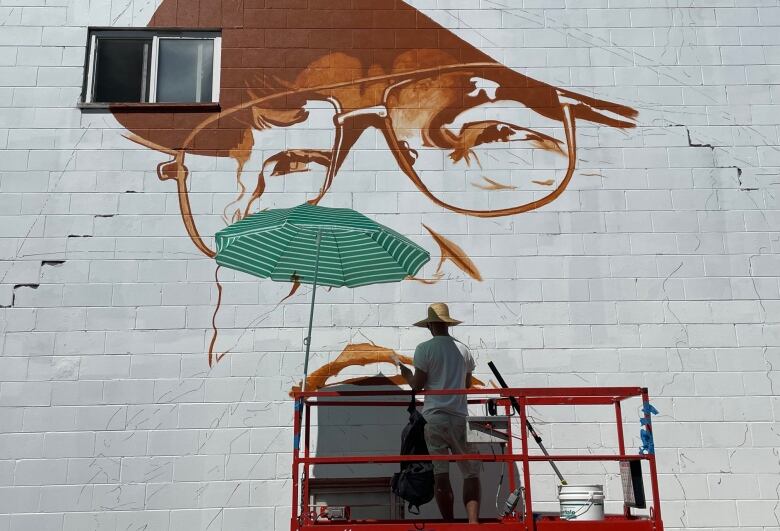 A painter in a wide-brimmed hat stands on a painter's platform. The beginnings of a painting, depicting a man with glasses, are visible on the wall in front of him.