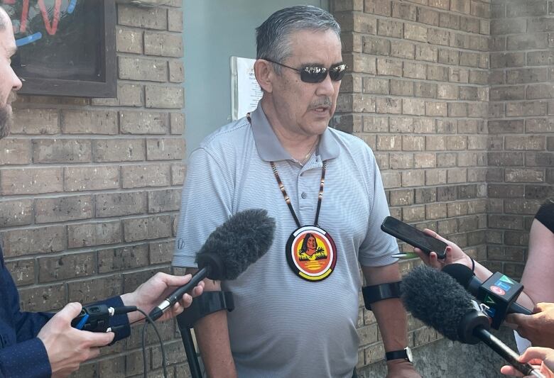 A man with grey hair, wearing dark sunglasses and a grey polo shirt speaks to reporters. 