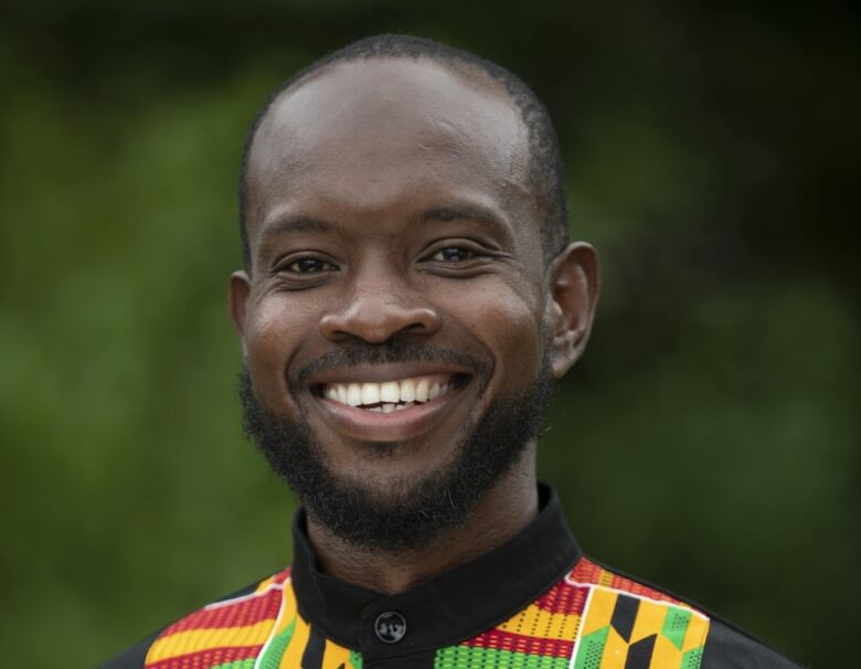 Photo of 2022 Sobey Art Award nominee Tyshan Wright. Pictured in medium closeup in a colour photo, Tyshan is a young Black man with very short hair and a close-cropped beard. He wears a button-down shirt made of kente cloth. He beams at the camera and stands in front of a lush green backdrop.