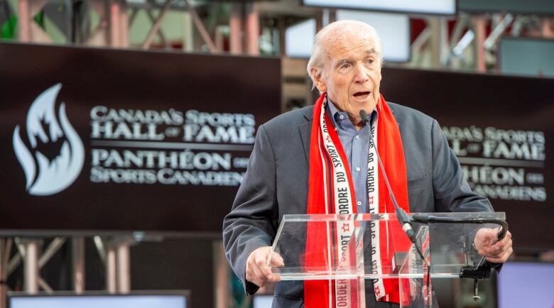 Doug Mitchell stands at a clear podium to speak as he's inducted into Canada's Sports Hall of Fame in Toronto on Oct. 23, 2019. He wears a red scarf that reads Order of Sport, as he and the other 2019 inductees were the first to be presented with an Order of Sport Award.