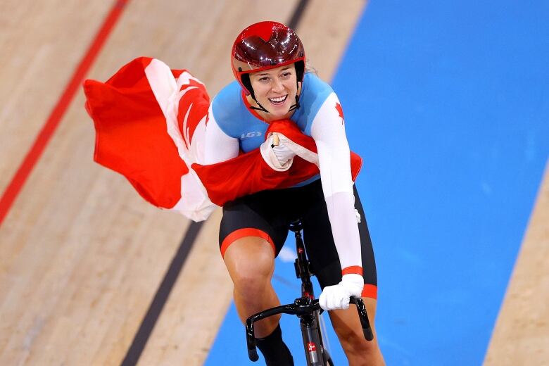 A smiling female cyclist rides a bike on an indoor track, clutching a Canadian flag to her chest.