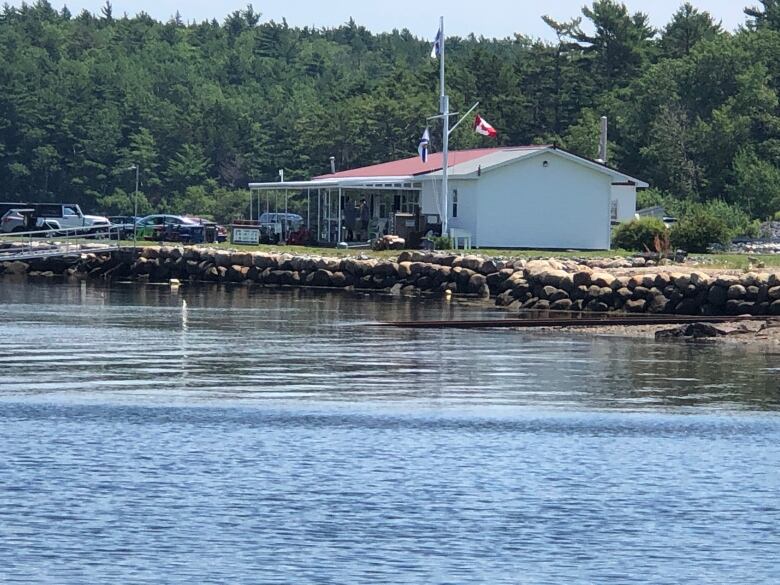 Blue water laps against the shore in front of a small white building backed by lush green forest. 